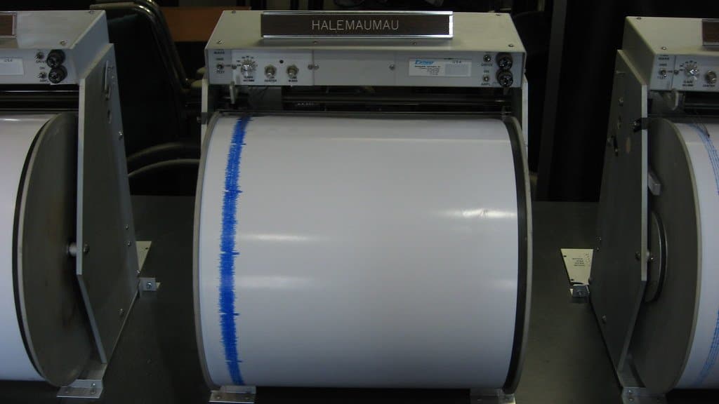 Three analogue paper-drum seismograph recorders in a row on a dark bench in a museum display; the central recorder carries an engraved HALEMAUMAU nameplate and its white paper drum shows a single ragged blue ink trace