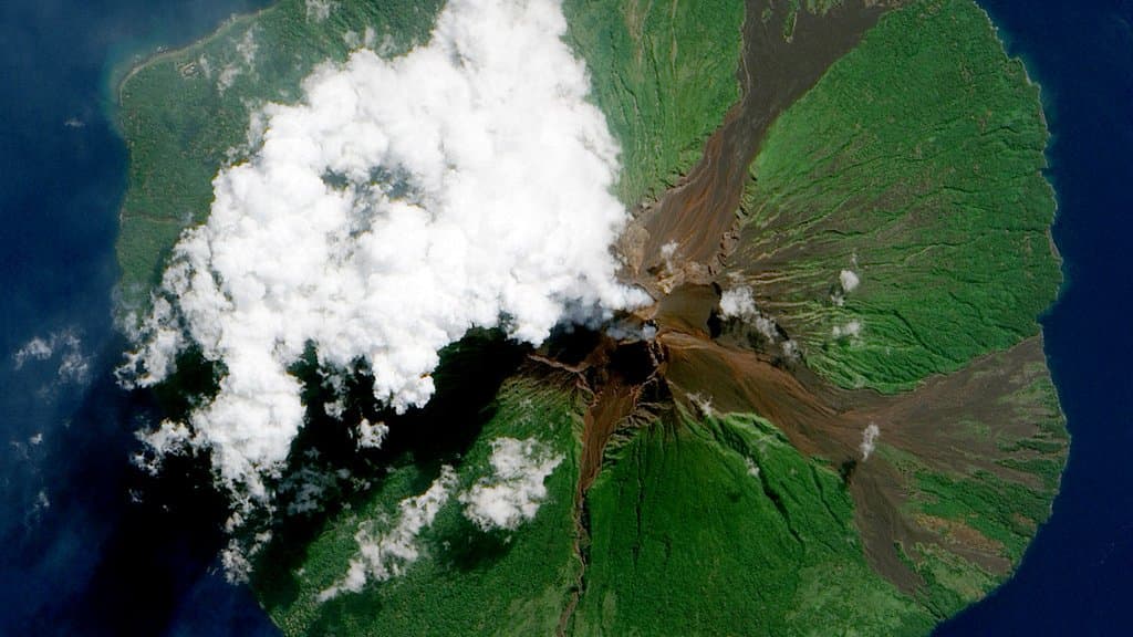 A satellite view of an erupting island volcano venting a plume over the surrounding ocean.