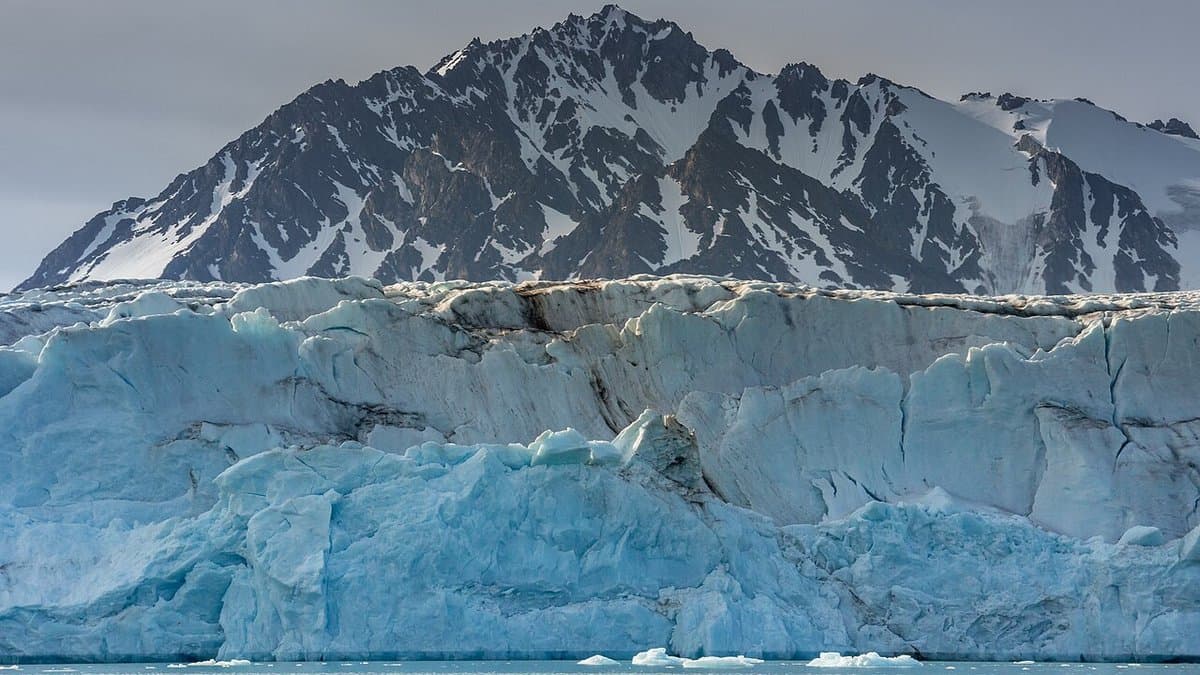 The blue ice cliff of a tidewater glacier meeting the sea in Svalbard, with a dark snow-streaked mountain rising behind it.