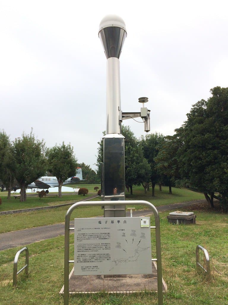 A stainless-steel GNSS pillar with a white antenna dome on top, standing in a grassed park beside an explanatory plaque reading denshi kijunten (electronic control point).
