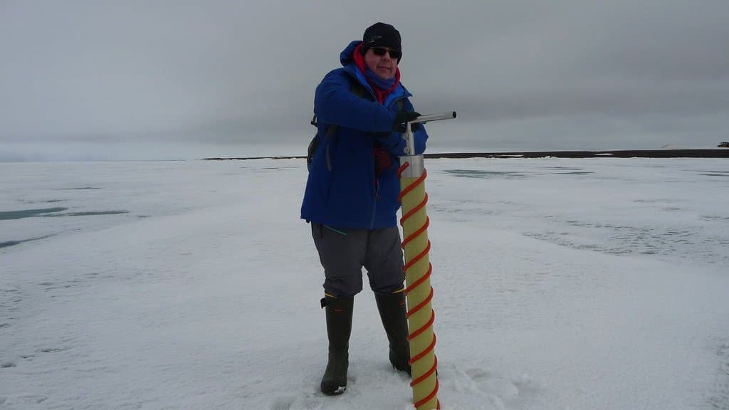 A researcher in a blue parka stands on flat sea ice holding a long spiral ice corer, with open water and a low shoreline behind