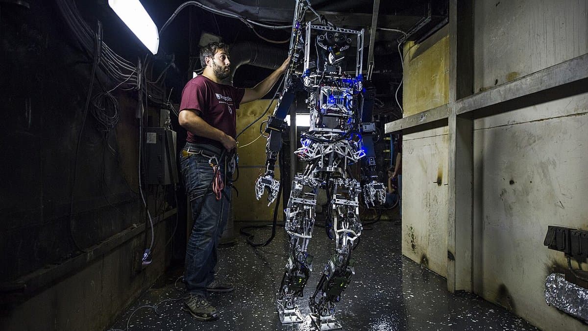 A researcher steadies a life-size bipedal humanoid robot by its frame during a trial in a narrow steel corridor