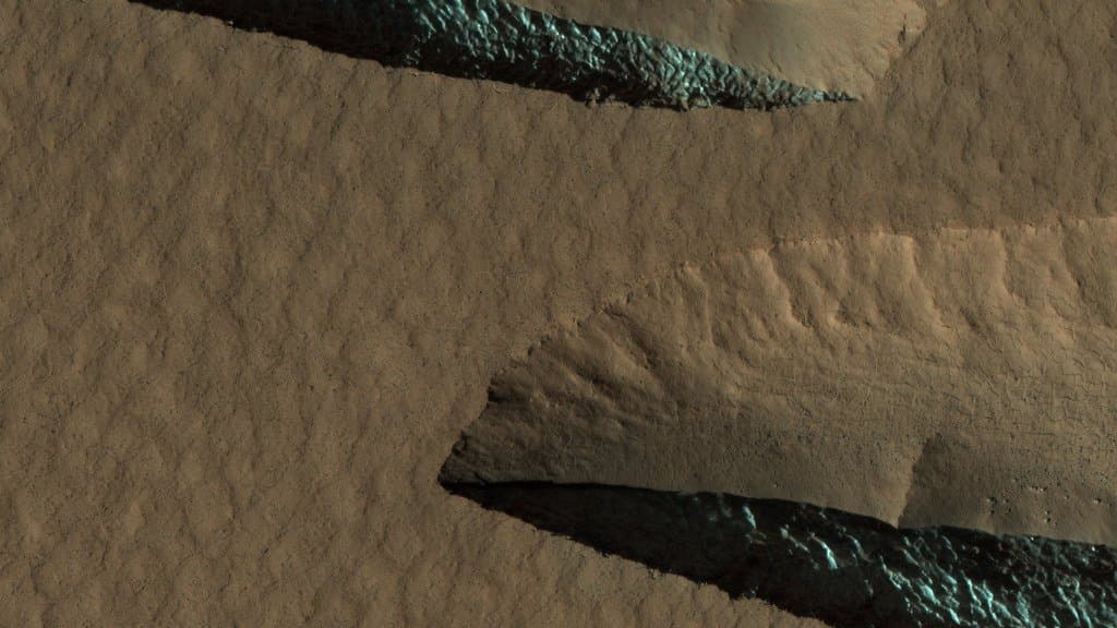 A HiRISE orbital image of the Martian mid-latitudes: rippled tan ground cut by two steep scarps whose faces expose bright blue-green ice.