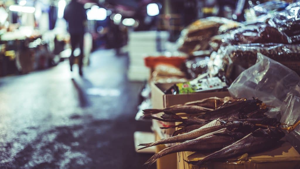 A narrow city market street at dusk, boxes of dried fish stacked on a stall in the foreground and further stalls and a lone shopper receding into the background.