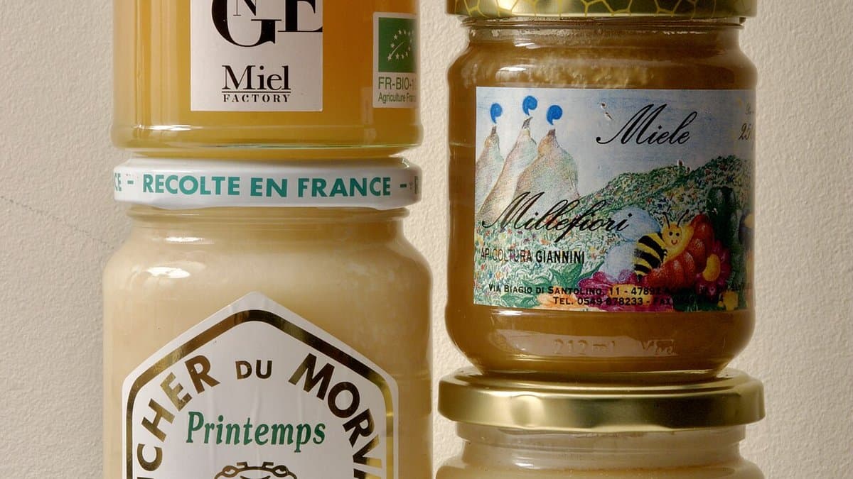A close view of stacked retail honey jars against a plain wall. Their printed labels are visible, including a green EU organic leaf certification mark and an FR-BIO code on one jar, a band reading recolte en France on another, and an illustrated Italian millefiori label on a third.