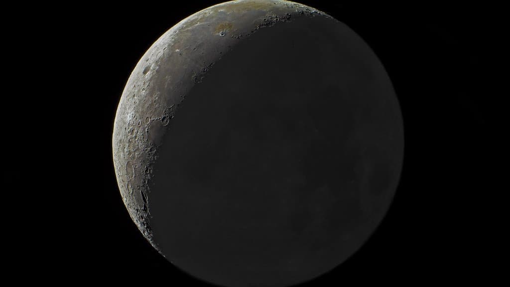 Telescopic photograph of the Moon: a narrow sunlit crescent along the left limb, with the rest of the near-side disc showing faintly in earthshine against a black sky.