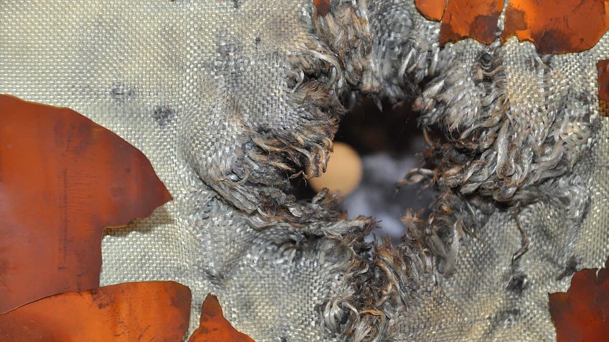 Extreme close-up photograph of spacecraft shielding after a hypervelocity impact test: a ragged hole punched through orange metal foil and layers of frayed grey ballistic fabric.