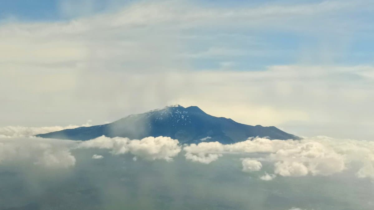 Mount Etna's broad snow-flecked summit ridge rising above a bank of low cloud, photographed from the air on a hazy day, with a thin wisp of white gas drifting from the summit crater area.