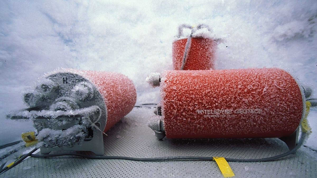 Three orange cylindrical instruments thickly furred with white frost crystals, one of them stencilled TELEDYNE GEOTECH, sitting with a grey finned sensor head and grey cables on a perforated metal base plate laid on snow under a flat white sky.