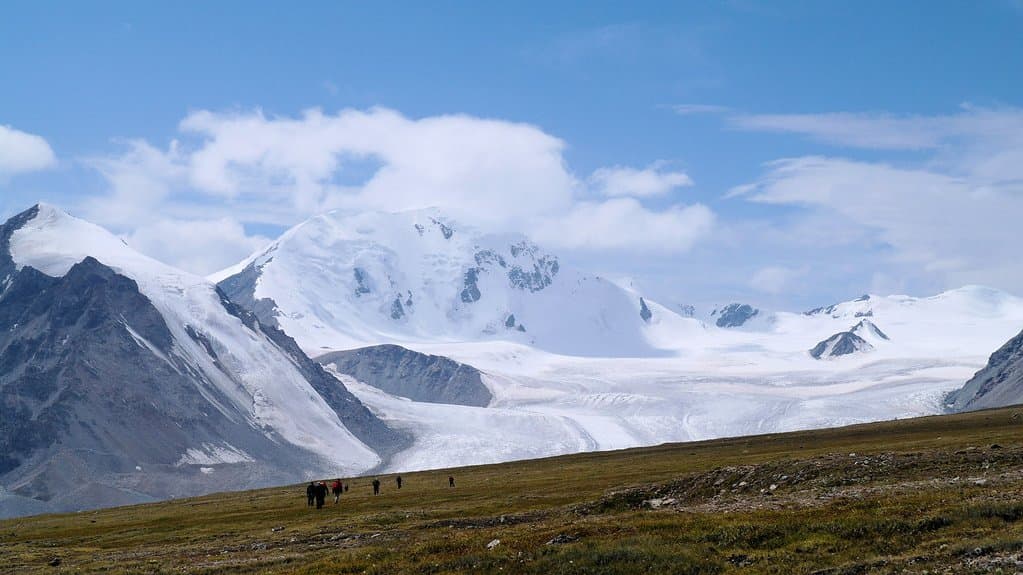 A wide alpine view: a broad snowfield and a debris-streaked glacier tongue spilling from a rounded snow-capped peak, with dark rock ridges either side, and in the foreground a long slope of dry gold-and-green tundra grassland where a line of half a dozen tiny walkers is crossing.