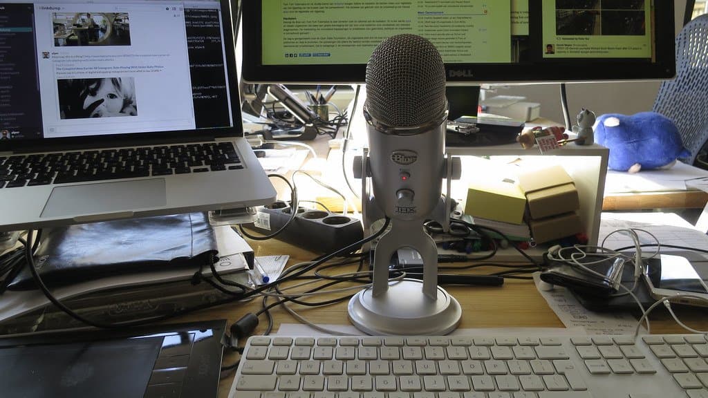 A cluttered desk with a large silver condenser microphone standing directly behind a computer keyboard, a laptop to the left and two monitors showing text behind it.