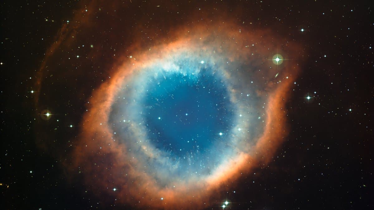 Wide-field telescope image of the Helix Nebula: a bright blue-green ring of glowing gas with a red outer rim, set against a dense star field.