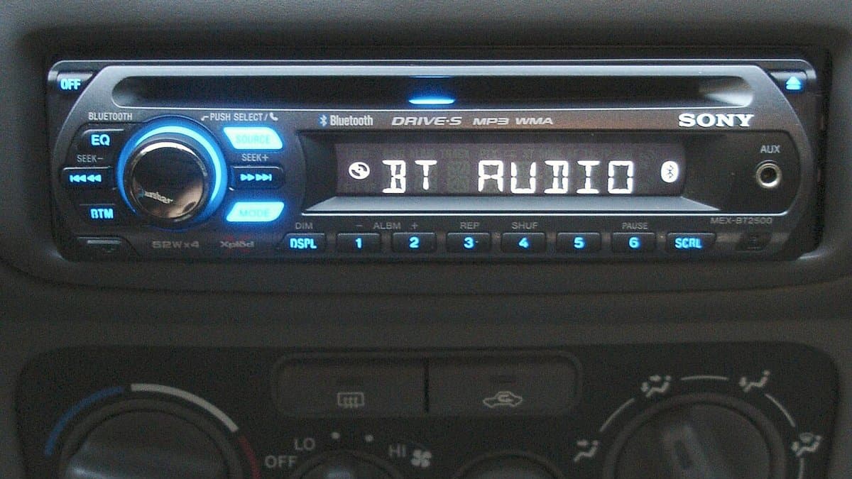 An illuminated aftermarket head unit fitted into a car dashboard, its display reading BT AUDIO above a row of preset buttons and heater controls.