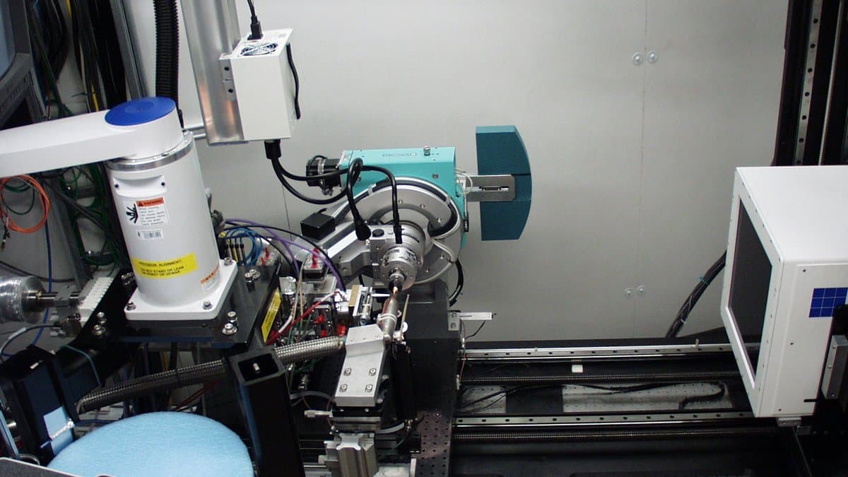 Inside an X-ray beamline experimental hutch: a sample goniometer and robotic sample arm in front of a detector mounted on rails, surrounded by motor stages, cabling and control boxes.