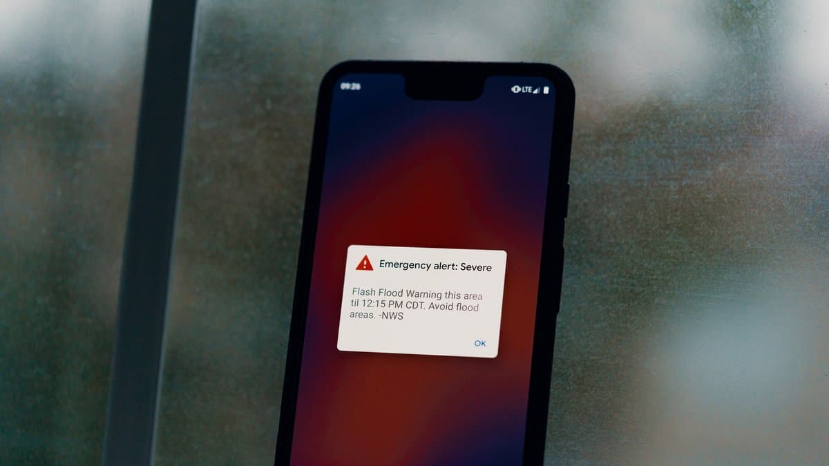 A smartphone held against a rain-streaked window, its screen showing a red-triangle ‘Emergency alert: Severe’ push notification.