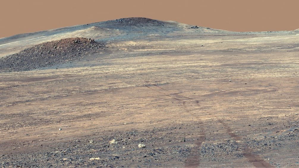 Wide view across a pale, dust-covered Martian slope, with a broad band of bright, light-toned rock running from the left of the frame to the right beneath darker, rubbly ridges, and faint rover wheel tracks curving down the slope in the foreground.