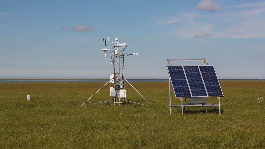 A solar-powered instrument mast standing on flat coastal tundra beside a bank of solar panels, under a pale Arctic sky