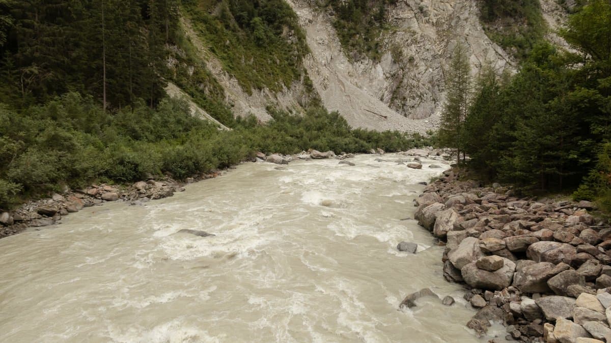 A turbulent, sediment-grey mountain river filling its channel between forested slopes, with boulder bank protection on one bank and a bare scree face rising behind