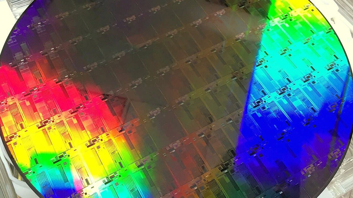 A silicon wafer patterned with rows of identical silicon-photonics dies, held under bright light so the etched optical circuits show as coloured diffraction.