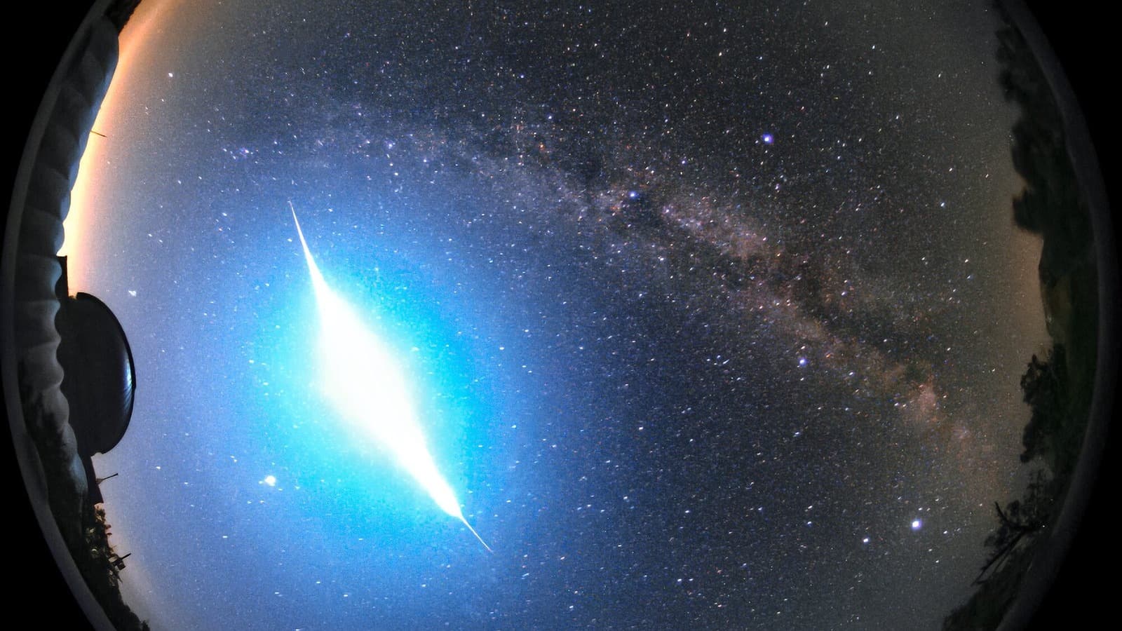 An all-sky camera frame showing a brilliant blue-white fireball blazing low in the night sky over Kitt Peak National Observatory, with the Milky Way arcing overhead and telescope domes silhouetted on the horizon
