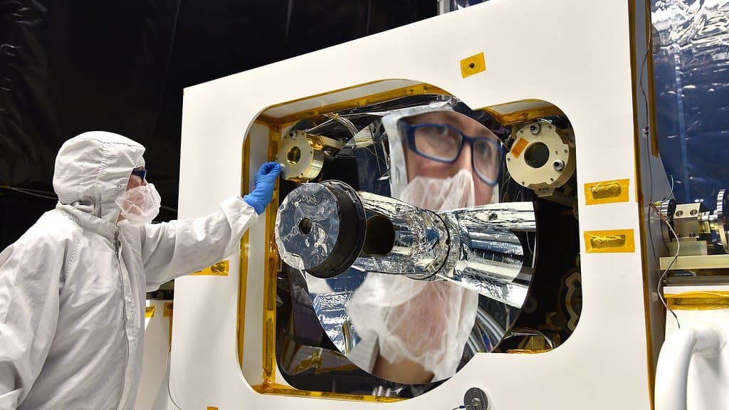 An engineer in a white clean-room suit reaches toward the large circular mirror of a space instrument, her face reflected in the polished surface