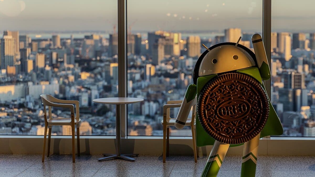 A statue of the Android robot mascot holding an Oreo biscuit, standing by a window above a city skyline at dusk