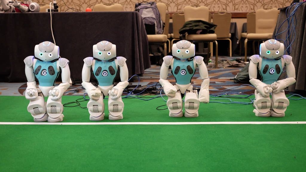 Four small humanoid Nao robots in matching blue team markings stand side by side at the edge of a green robot-football pitch, cables and venue furniture behind them.