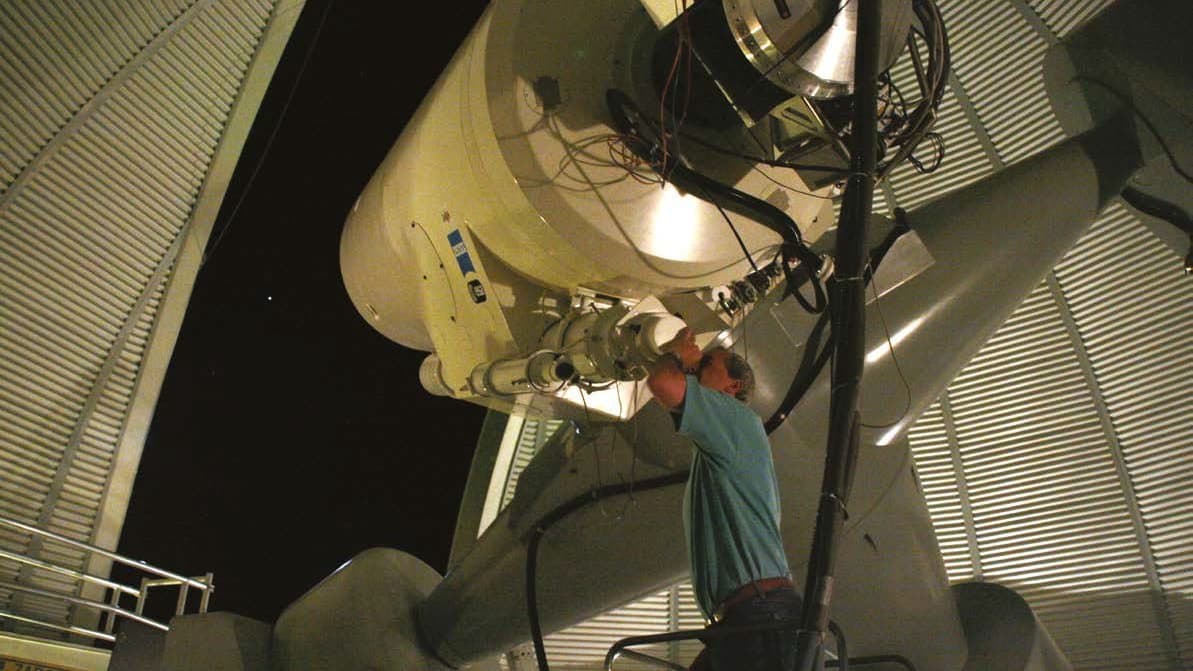 A man works at the eyepiece end of a large telescope inside an open observatory dome at night