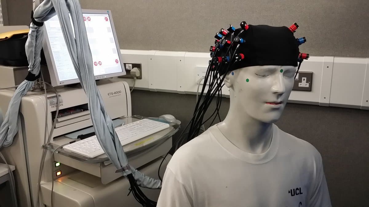 A mannequin head fitted with a functional near-infrared spectroscopy cap, with its optodes wired by black cables to a Hitachi ETG-4000 machine in a laboratory