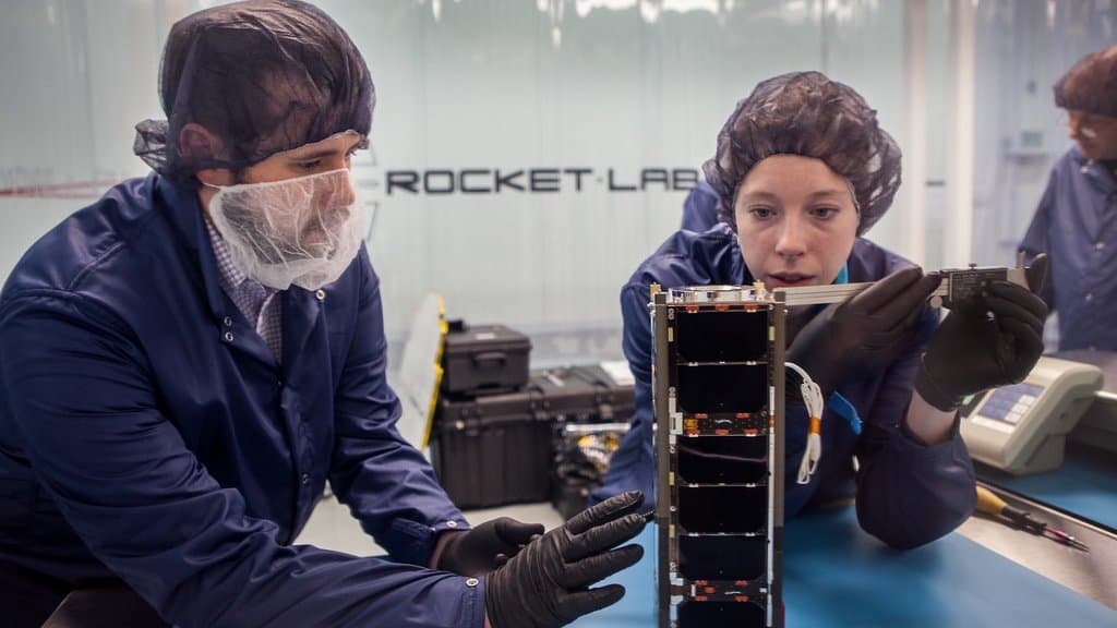 Two engineers in clean-room caps and gloves examine a small rectangular CubeSat on a workbench, one measuring it with calipers, under a Rocket Lab sign.