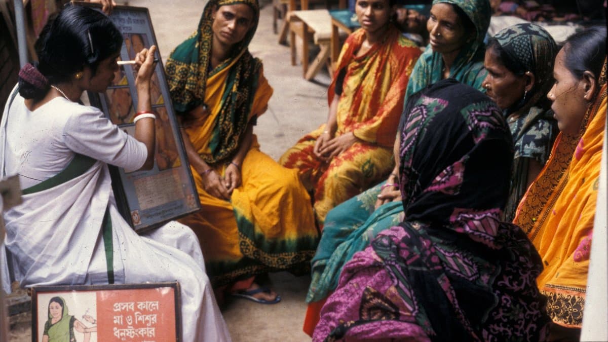 A health worker holds up an illustrated flipchart to a seated group of women in brightly coloured saris in an open passageway, with a Bengali-language poster about mother and newborn care propped on the ground beside her.