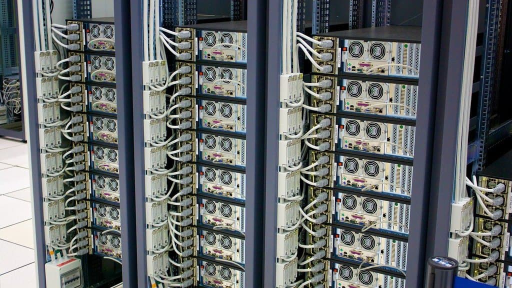 Rows of rack-mounted server units stacked floor to ceiling in a data centre aisle, each unit trailing pale network and power cables.