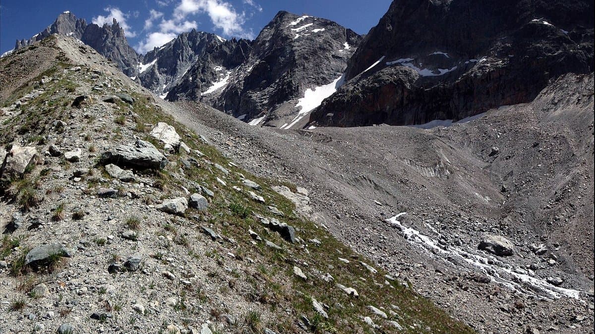 The stony, debris-covered tongue of a glacier filling a high valley below the rock walls and snowfields of the Écrins massif, seen across a moraine slope.