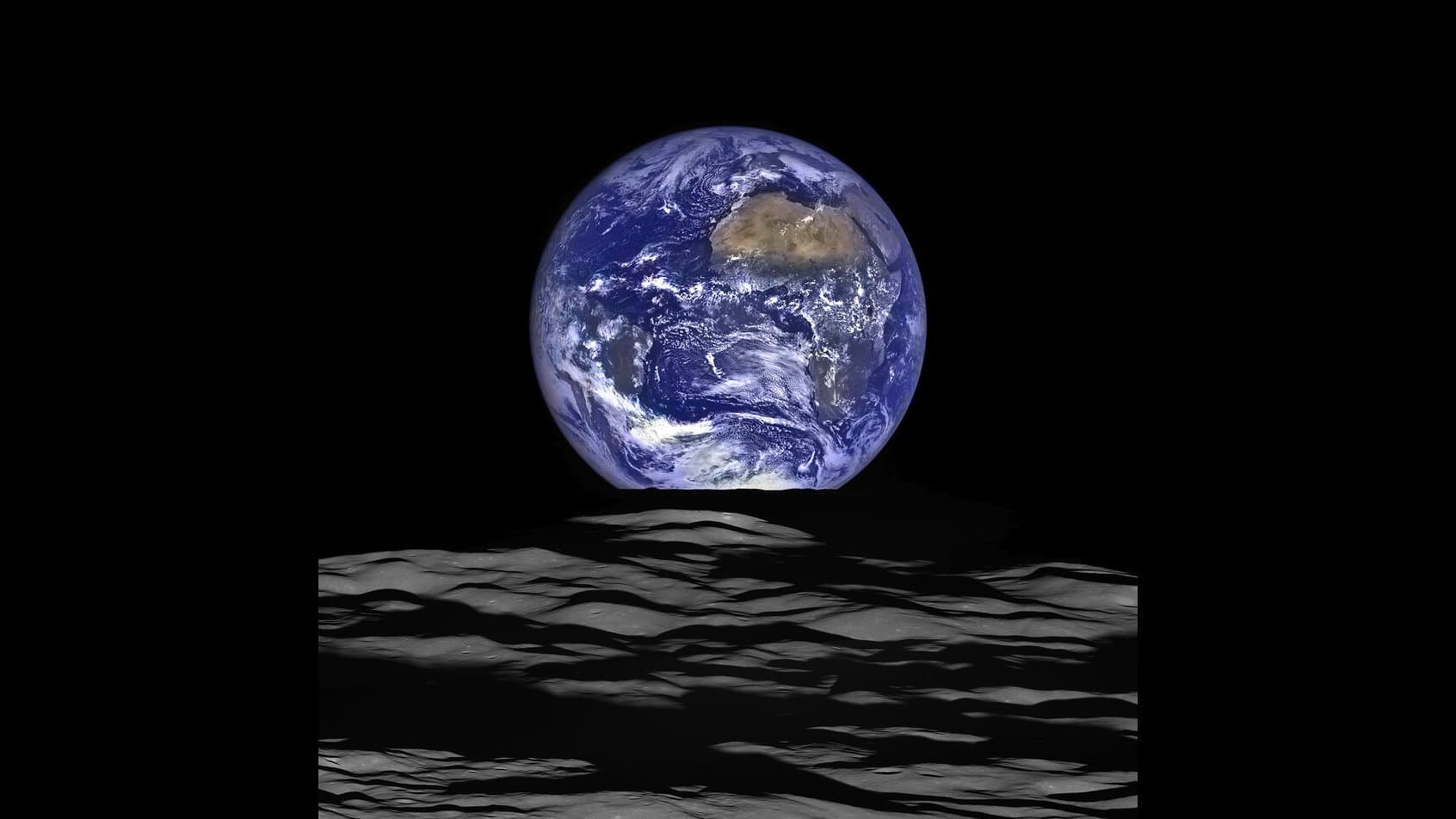 The whole disc of Earth, blue oceans and white cloud with the Sahara visible, hanging in black space above the grey, cratered horizon of the Moon, photographed from lunar orbit.
