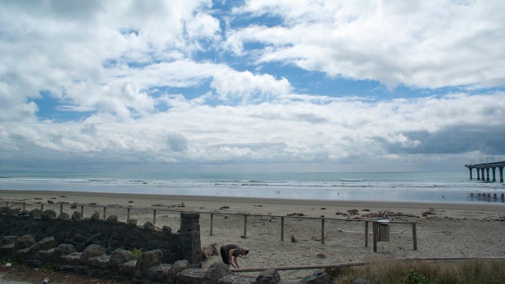 A wide sandy beach under broken cloud, the waterline running across the middle distance and a long pier reaching out to sea on the right.