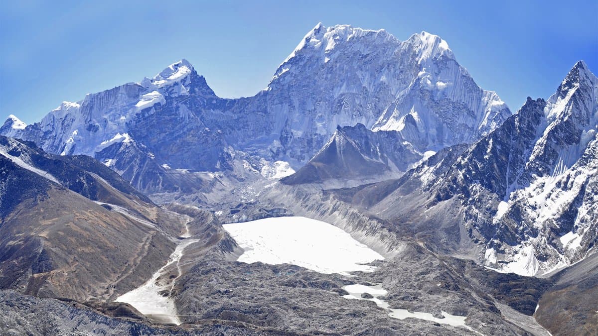 A high-mountain glacial lake held behind a broad ridge of rock and debris, with snow peaks rising behind it and a rubble-covered glacier valley in front.