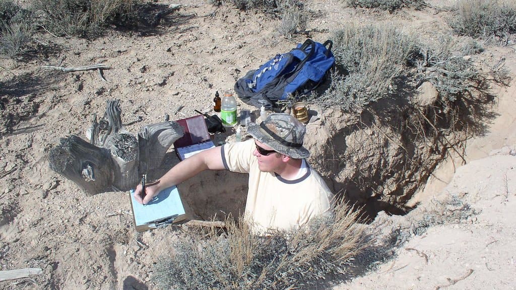 A soil scientist standing waist-deep in a freshly dug soil pit, writing notes on a clipboard beside sampling gear.