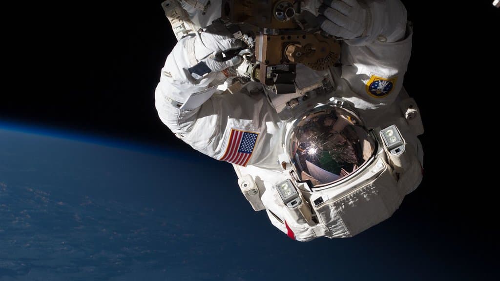 A spacesuited astronaut outside the International Space Station, gold visor reflecting the station, with the curve of Earth below.