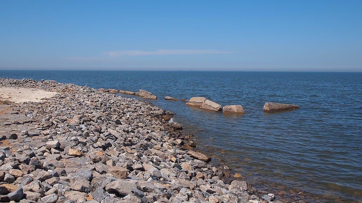 A shoreline of loose grey boulders meeting the calm shallow water of the Bohai Sea under a clear sky.