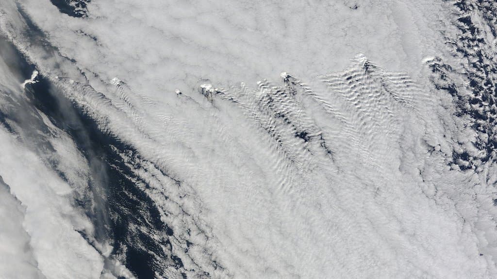 A satellite view of a thick white deck of low marine cloud over dark ocean, rippled into wave patterns downwind of a chain of islands.