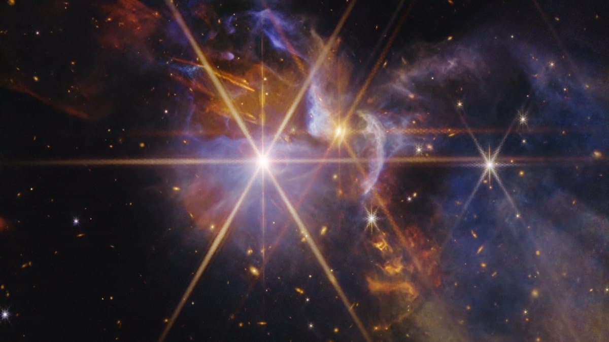 A James Webb Space Telescope near-infrared image of the young star system FS Tau: a brilliant star with long diffraction spikes, surrounded by orange and blue clouds of gas and dust and a curved outflow arc.