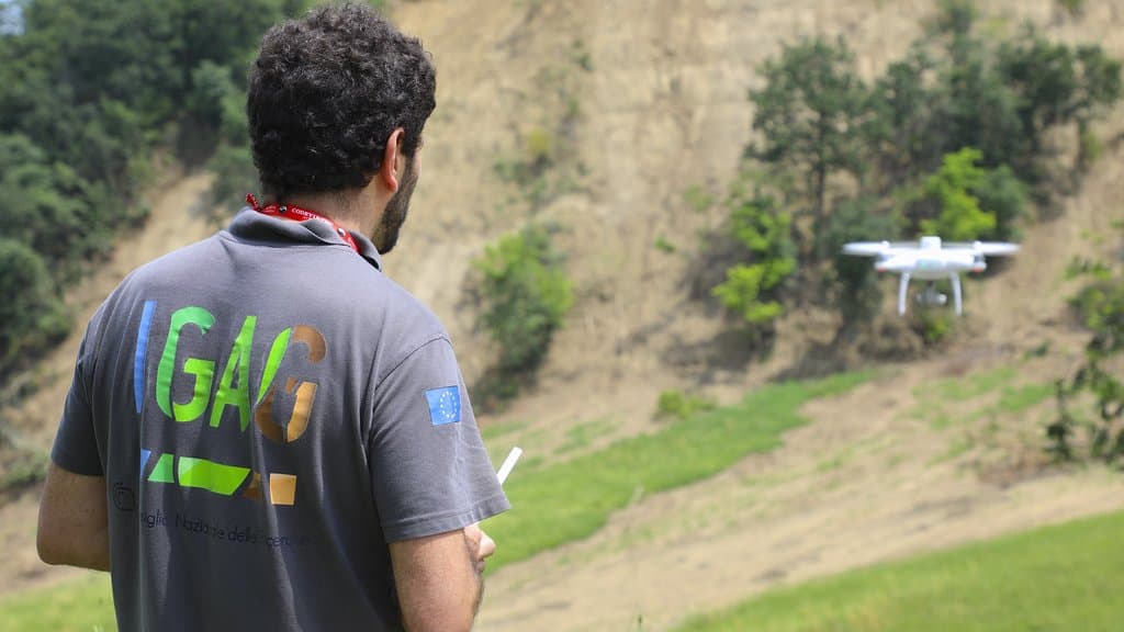 A researcher in an IGAG shirt stands with his back to the camera, flying a small white quadcopter over a bare landslide scar cut into a green Italian hillside.