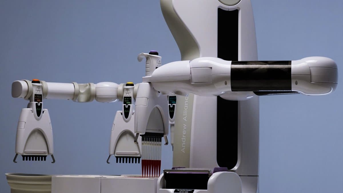 A white robotic arm reaching over a rack of electronic pipettes on a laboratory bench