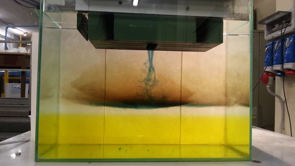 A glass tank of layered fluid standing on a laboratory bench, a metal plate suspended across the top, and a plume of blue dye sinking through an orange layer towards a yellow one below.