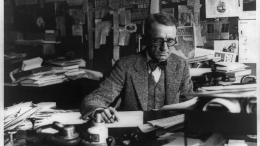 Black-and-white archival photograph of a man in a tweed suit and bow tie at a desk piled with manuscripts and loose pages, newspaper clippings pinned to the wall behind him.