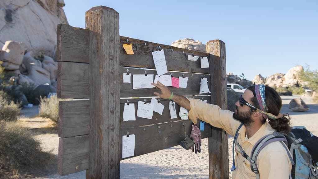 A hiker reaching up to a wooden public message board hung with handwritten notes