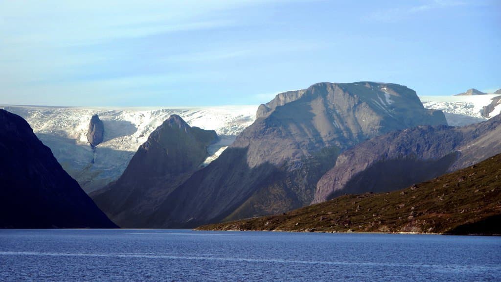 A steep-sided South Greenland fjord with dark water in the foreground and glaciers spilling between bare rock mountains from an icecap behind.
