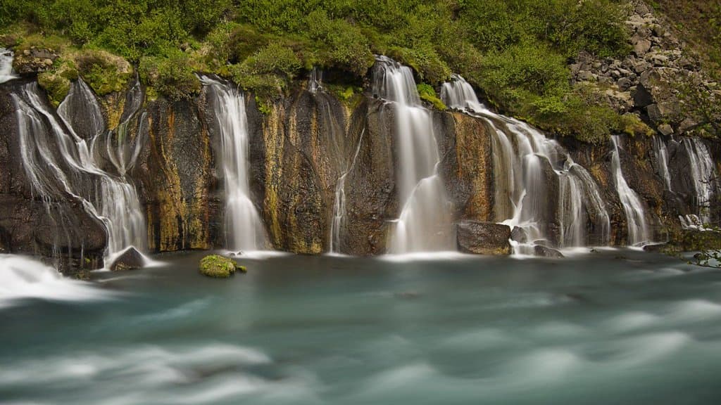 A long line of small waterfalls emerging from beneath a mossy lava field and spilling into a wide turquoise river.