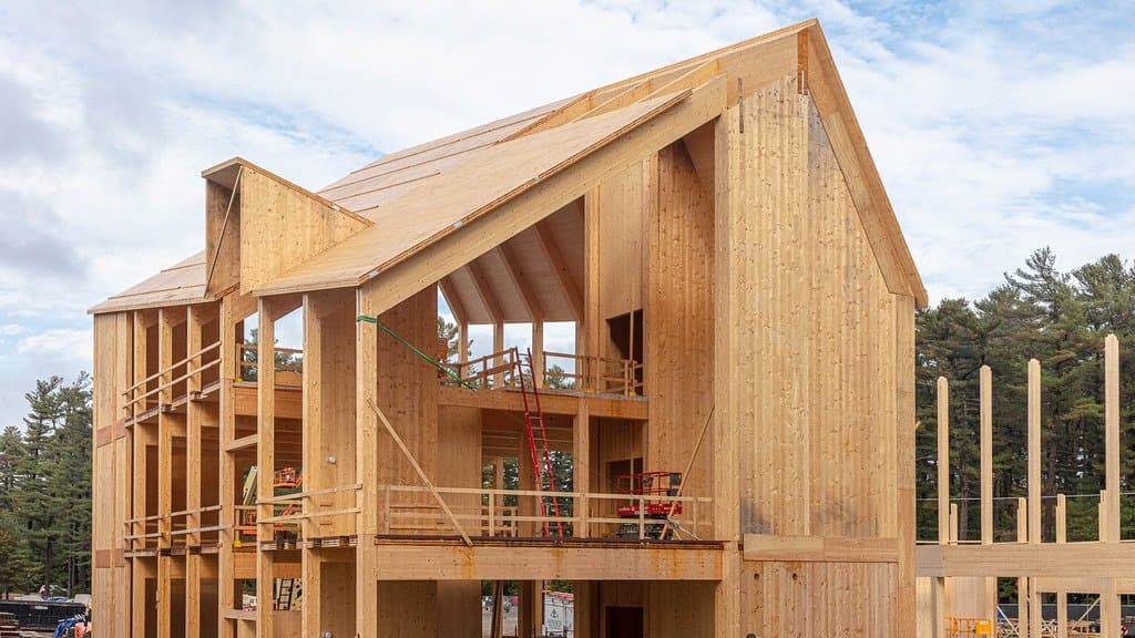 A three-storey building under construction with its exposed frame, floors and roof built entirely from pale engineered timber, with excavators parked in front.
