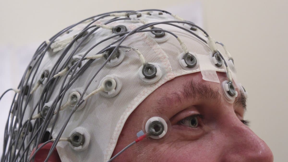 A person wearing a fitted EEG cap covered in numbered electrode sockets and trailing recording leads